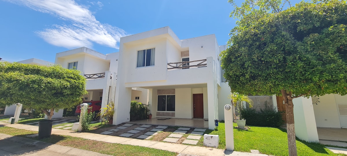 A modern two-story home is presented with smooth white walls and a welcoming entrance. Neatly arranged stone pathways lead to the front door, framed by green shrubs and trees. Large windows allow natural light to enter, complementing the bright exterior.