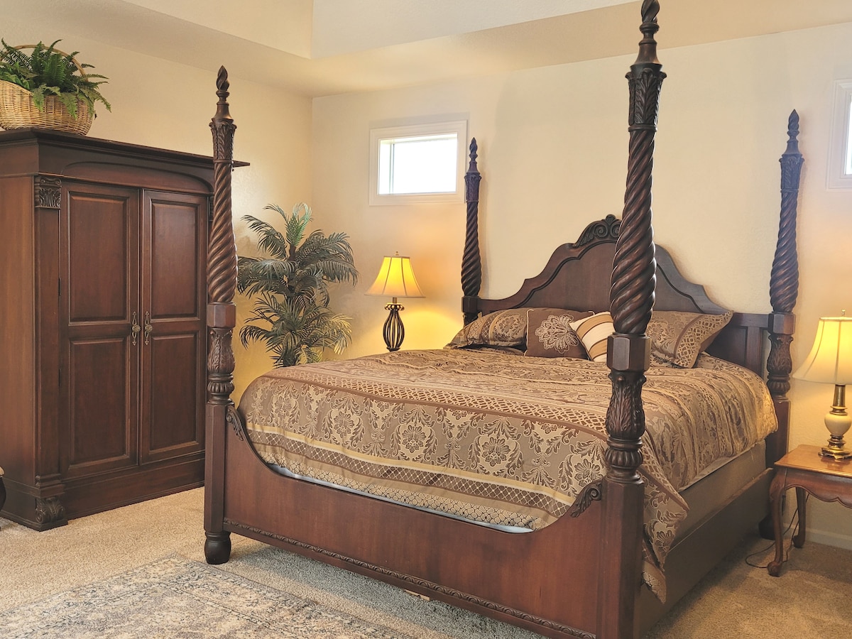 A spacious master bedroom features a four-poster bed adorned with a patterned bedspread. An ornate wooden wardrobe stands adjacent, accompanied by a decorative lamp and a small side table. Natural light enters through high windows, illuminating the calming space.