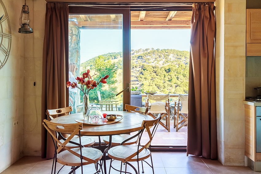 A round dining table is set with a vase of flowers and snack items, surrounded by four wooden chairs. Large glass doors open to a view of the mountains and outdoor seating area, while warm curtains frame the entrance.