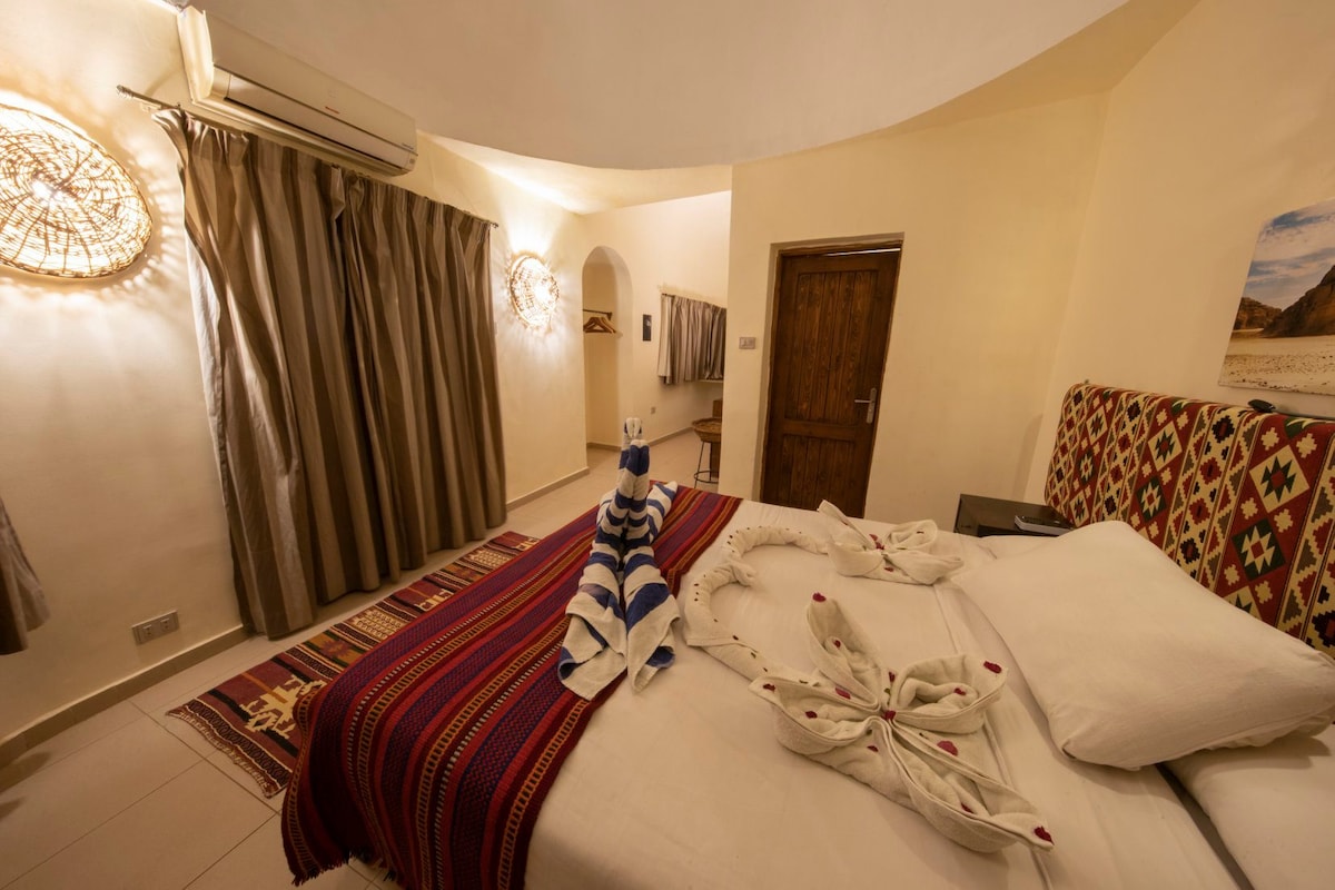 The room features a spacious bed adorned with neatly arranged towels, styled to resemble a playful figure. Light beige walls are complemented by decorative woven light fixtures and curtains, while a patterned rug adds warmth to the space. A wooden door leads to another area.