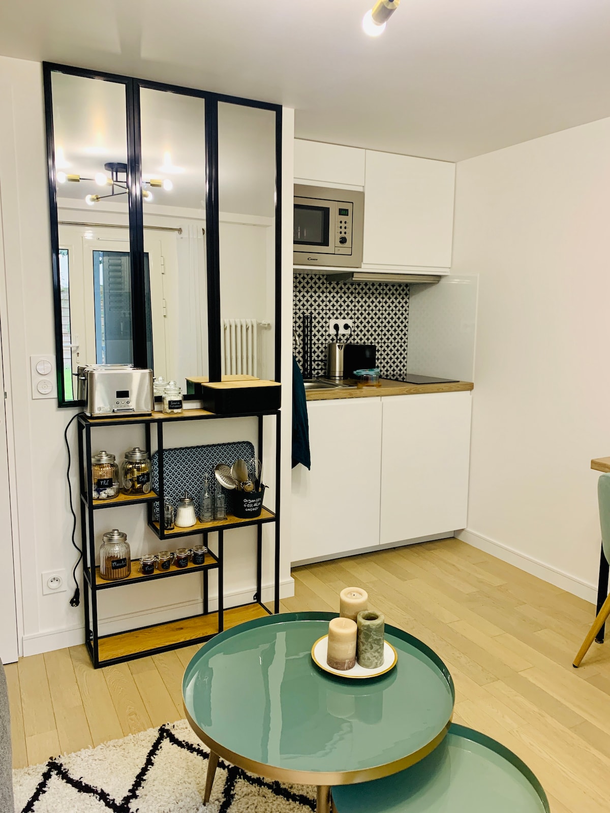 A compact kitchenette is equipped with modern appliances, including a microwave and a stovetop. A stylish mirrored cabinet holds kitchenware, while a round coffee table with decorative candles sits in the foreground, complementing the overall clean and functional design.