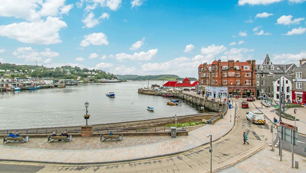 A scenic view of the harbour in Oban, highlighting the water's calm surface and activity from boats. Surrounding buildings and shops line the waterfront with a vibrant mix of architecture. People are seen strolling along the promenade, reflecting the lively atmosphere of the area.