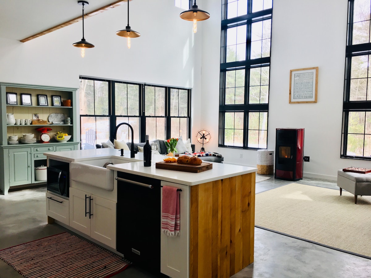 A spacious kitchen is complemented by large windows that allow natural light to fill the area. An island with a countertop of mixed materials serves as a central gathering spot. A colorful rug lies on the concrete floor, while various kitchenware is neatly arranged on shelves.