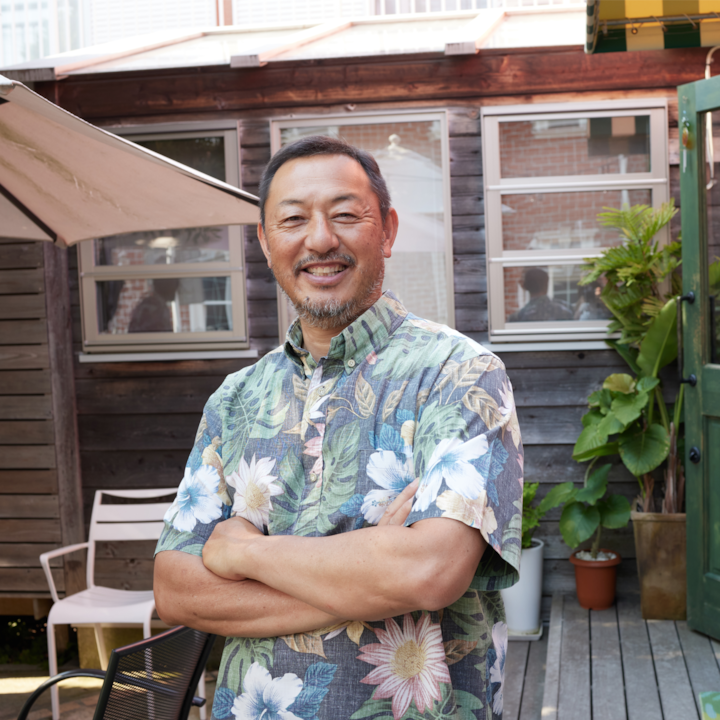 A person with a goatee in a Hawaiian shirt smiles on a sunny wooden deck with plants and a patio umbrella.