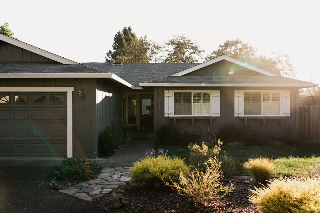 The front exterior of Siesta Cottage features a welcoming façade with light grey siding and white shutters. A stone pathway leads through the garden, where various shrubs and plants are arranged. Morning light casts a soft glow on the home.