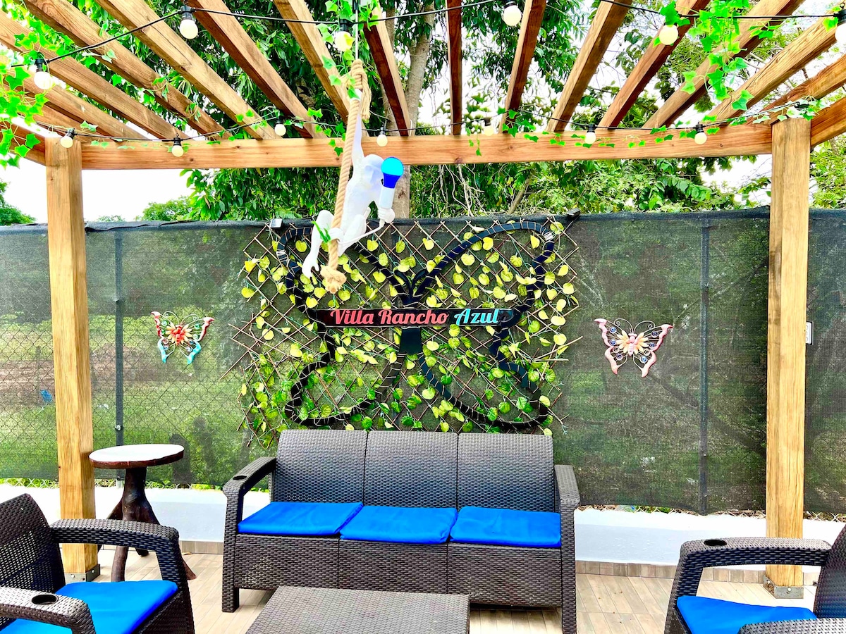 A shaded outdoor seating area is showcased with a wooden pergola adorned with greenery. Comfortable dark furniture with blue cushions is arranged, creating a welcoming space. Decorative butterflies and a sign reading 'Villa Rancho Azul' add character to the backdrop.