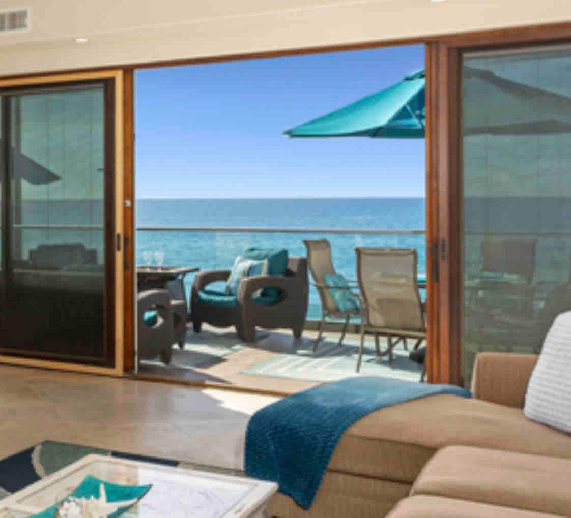 Large glass sliding doors open to a private balcony, showcasing a panoramic ocean view. Outdoor seating accommodates several guests, complemented by a sunshade umbrella. The interior living space features a comfortable sofa and decorative elements, creating a welcoming environment with natural light.