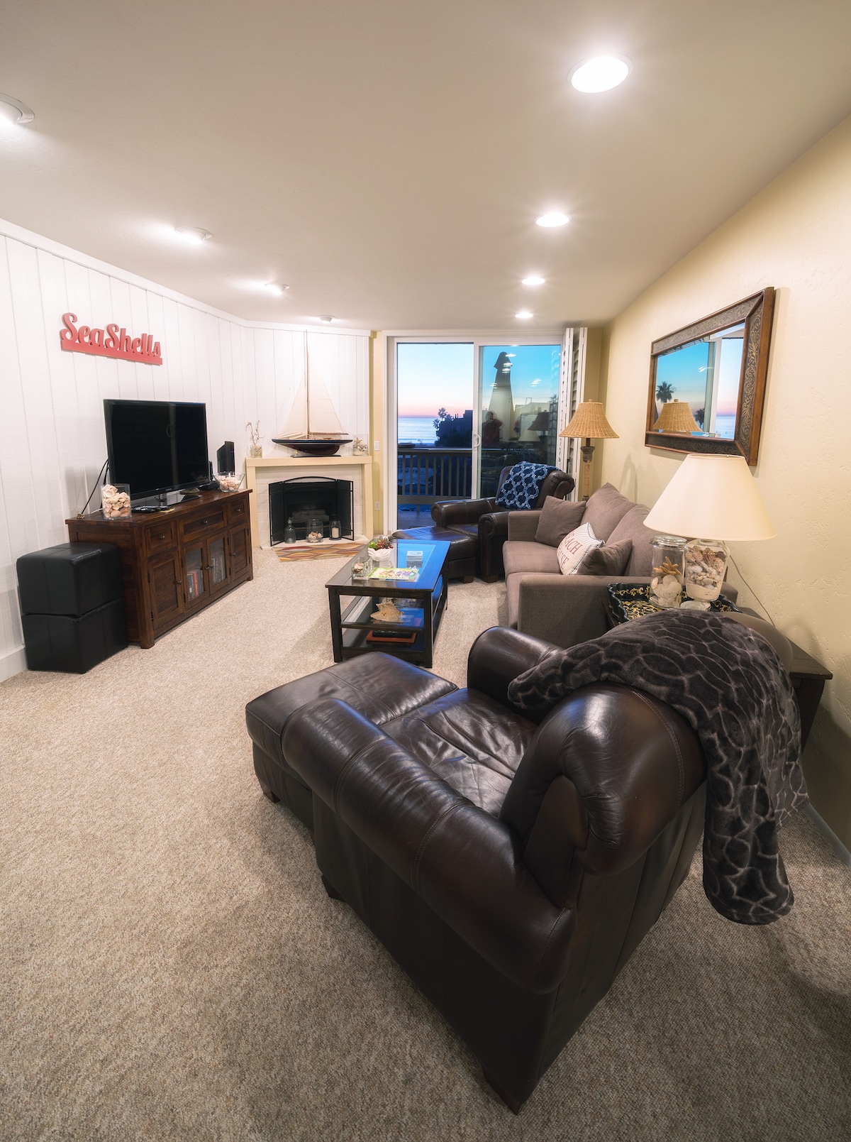 A cozy living area features two comfortable armchairs and a plush sofa, arranged around a coffee table. A flat-screen TV is situated on a wooden console, while a lamp adds gentle illumination. A large window offers a view of the ocean, with soft colors of twilight in the background.