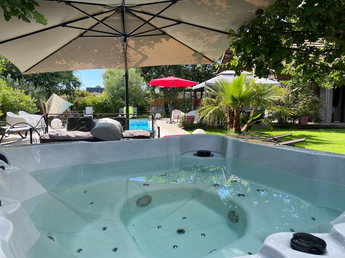 A spa is situated beneath a large shaded umbrella, surrounded by lush greenery and a well-kept garden. Relaxing seating areas, including loungers and a covered terrace, are visible in the background, providing a serene outdoor space to unwind.