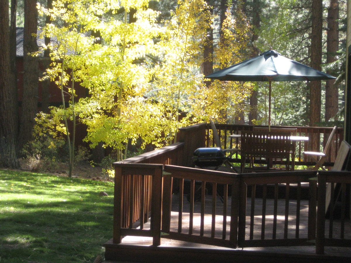A spacious wooden deck is surrounded by trees with bright yellow leaves. A green umbrella provides shade over a wooden dining set, while a grill is positioned nearby. The well-maintained lawn features lush green grass, enhancing the outdoor relaxation area.