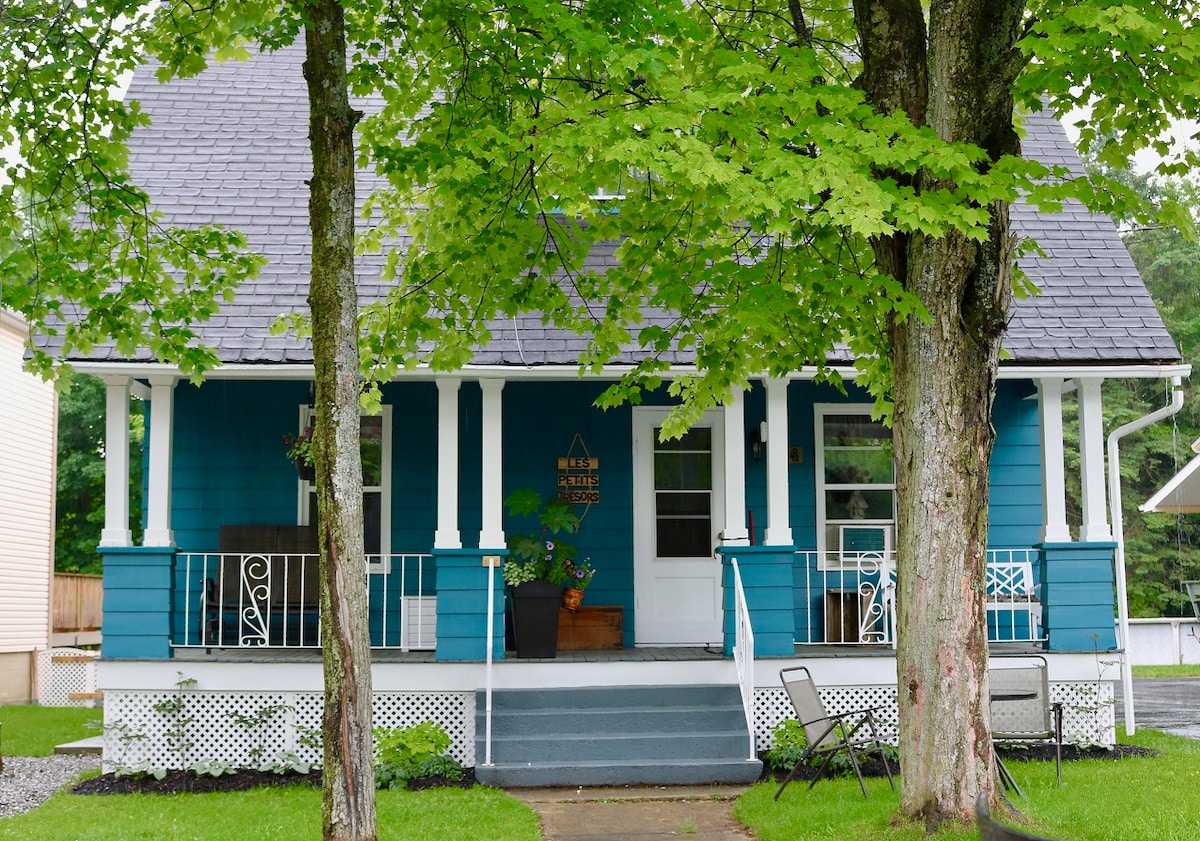 A charming blue house is framed by tall green trees, showcasing a welcoming front porch with white railings and chairs. The entrance features a decorative sign and potted plants, enhancing the inviting character of the property.