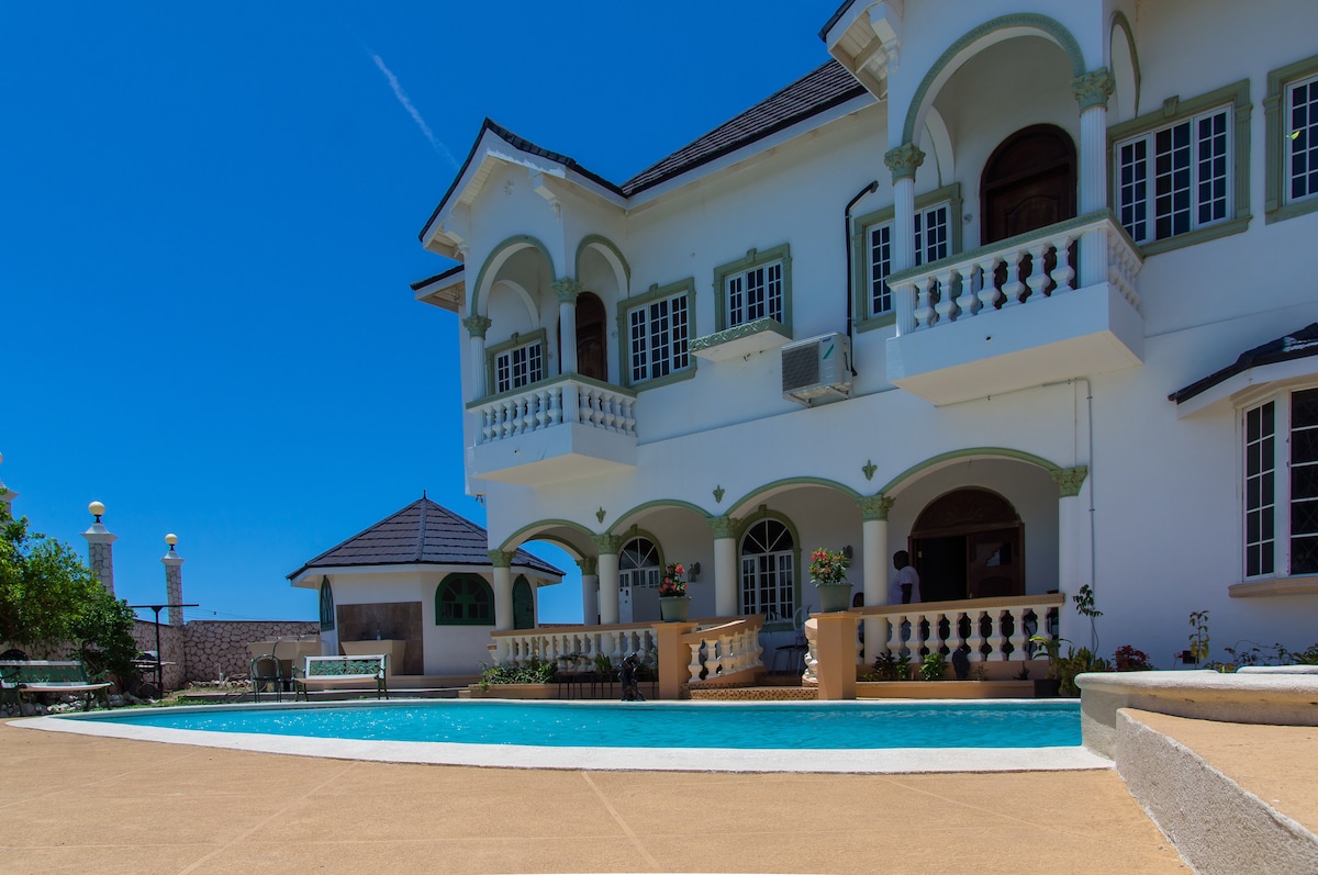 A large two-story house features arched balconies and detailed architectural elements. A swimming pool is situated in front, surrounded by a beige deck. The clear blue sky above adds to the bright ambiance, highlighting the home's inviting outdoor space.