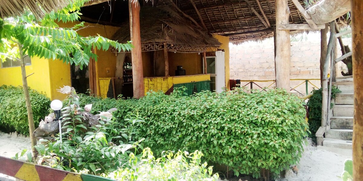 A vibrant garden area is surrounded by low, lush greenery and a sandy path. A rustic, yellow structure is visible in the background, featuring a thatched roof and large wooden beams. Steps lead to a raised area, enhancing access to the building.