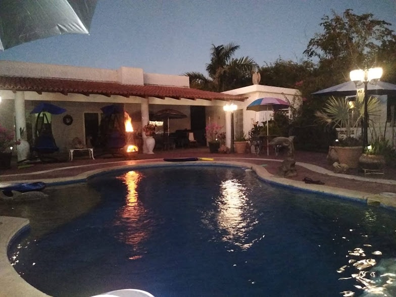 The image features a serene pool area illuminated by soft lighting as evening approaches. Surrounding greenery and various seating options, including colorful umbrellas and hanging chairs, create a welcoming outdoor space for relaxation and enjoyment.