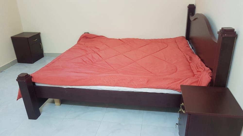 A spacious bed with a red blanket is situated in the center of the room, flanked by two dark wood nightstands. The floor features light-colored tiles, and the walls are painted in a neutral tone.