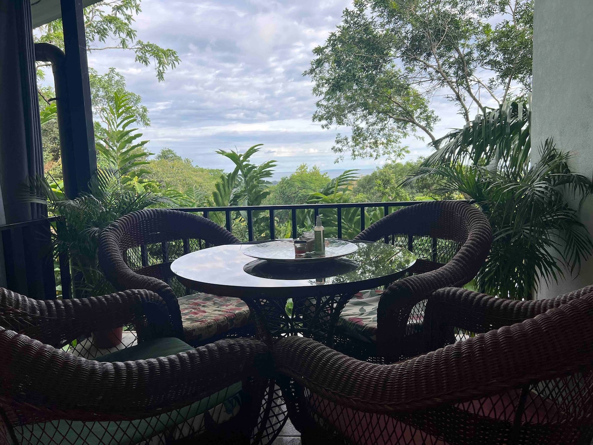 A terrace features a round table surrounded by four wicker chairs, accented with cushions. Lush greenery and tropical plants frame the view, which overlooks a distant ocean and cloudy sky, creating a serene outdoor space for relaxation.