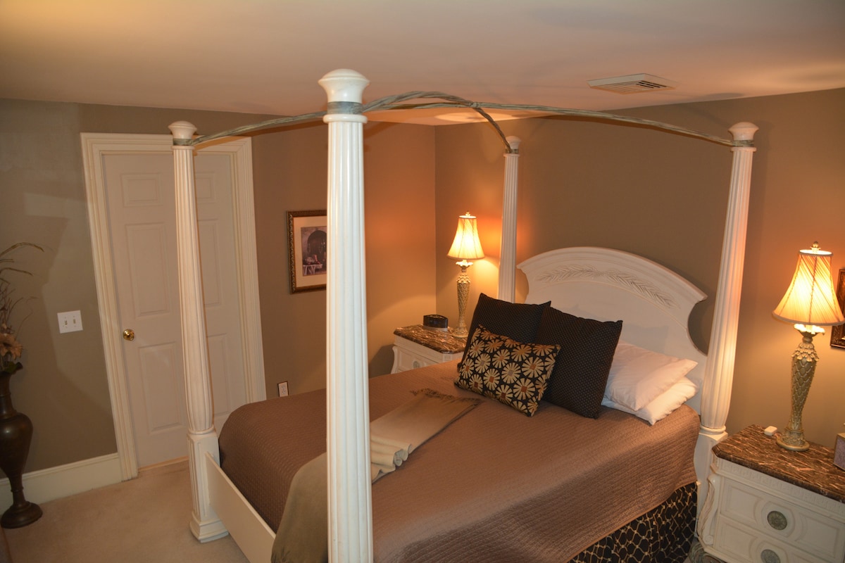 A cozy bedroom features a four-poster queen bed adorned with neutral bedding and decorative pillows. Two bedside lamps emit a warm glow, complementing the earthy wall tones. A framed picture hangs on the wall, and nightstands are positioned on either side of the bed.