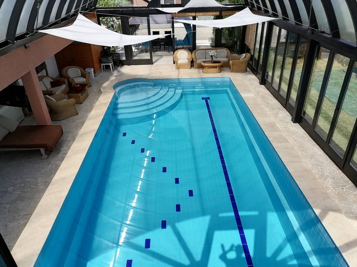 An indoor swimming pool is surrounded by large glass panels, offering ample natural light. White fabric drapes create a soft contrast above the water's surface, which features a pattern of blue tiles. Comfortable seating areas are visible in the background.