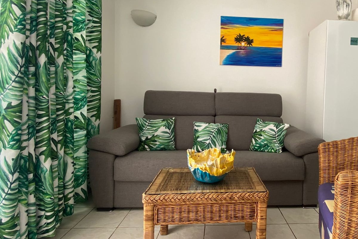 A living area features a gray sofa adorned with cushions that reflect green foliage. A wicker coffee table holds a colorful bowl. Brightly patterned curtains complement a wall art piece showcasing a tropical scene. Natural light fills the space, enhancing its relaxed atmosphere.