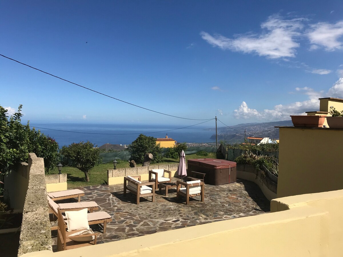 An outdoor terrace features a hot tub and several lounge chairs arranged on a stone covering. A clear view of the ocean is visible in the background, along with lush greenery and distant hills. Clear blue skies create a serene atmosphere.