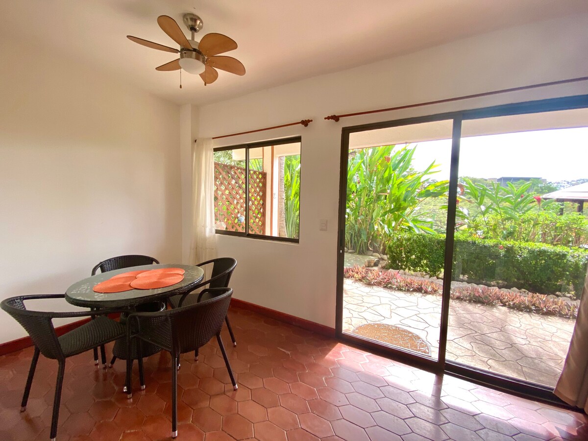 A dining area features a round table surrounded by four black wicker chairs. Natural light fills the space through large sliding glass doors, which open to a patio and a garden with lush green foliage visible outside.