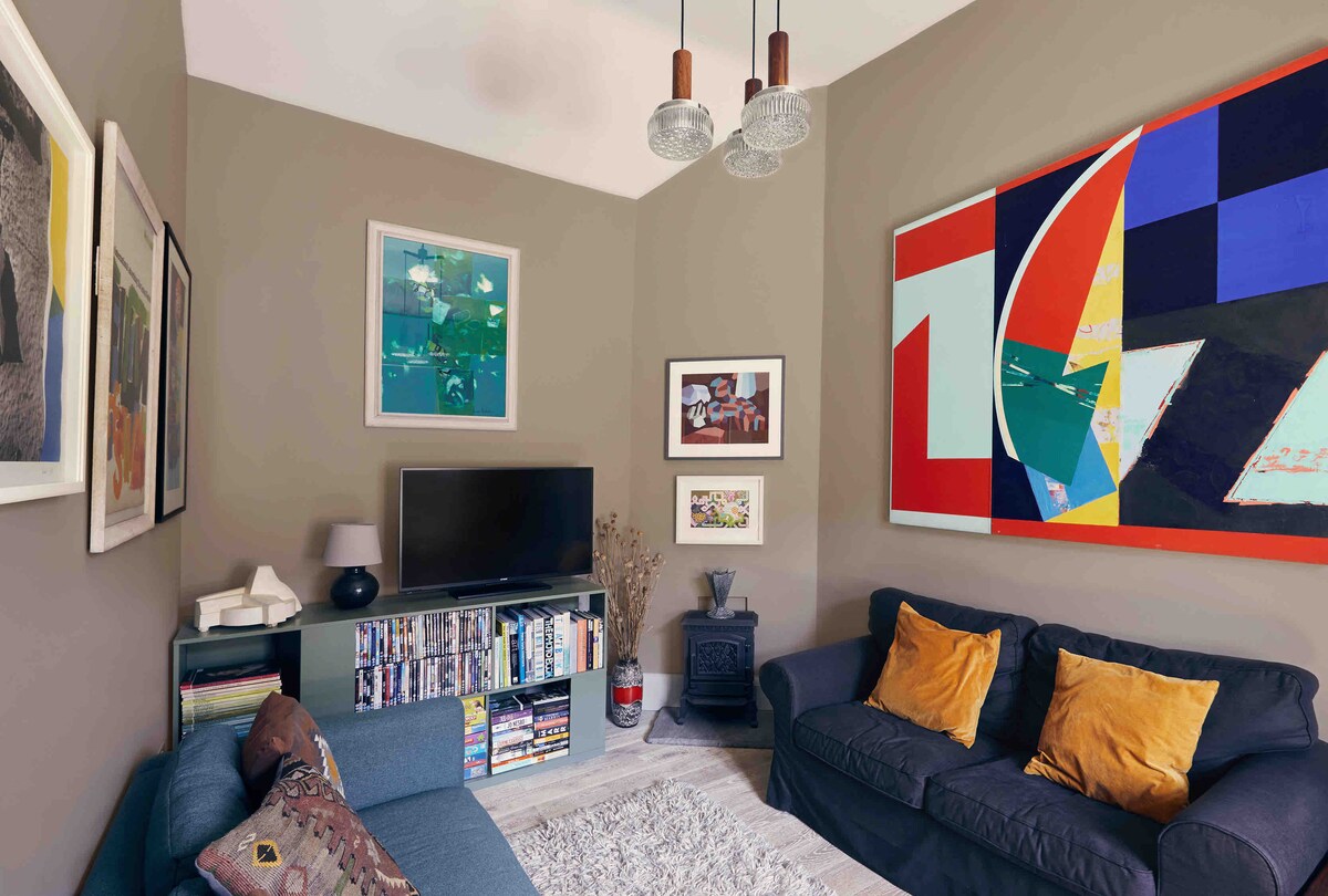 A comfortable sitting area features a blue couch and a dark sofa, both adorned with orange cushions. A colorful abstract painting dominates one wall, while a large TV is positioned on a low shelf beside a collection of books. An electric stove adds warmth to the space.