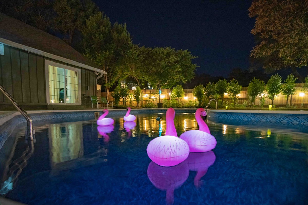 The tranquil pool is illuminated by soft lighting, reflecting vibrant pink from three inflatable flamingos. Lush greenery and a privacy fence frame the space, while gentle illumination from garden lights adds to the serene evening ambiance.