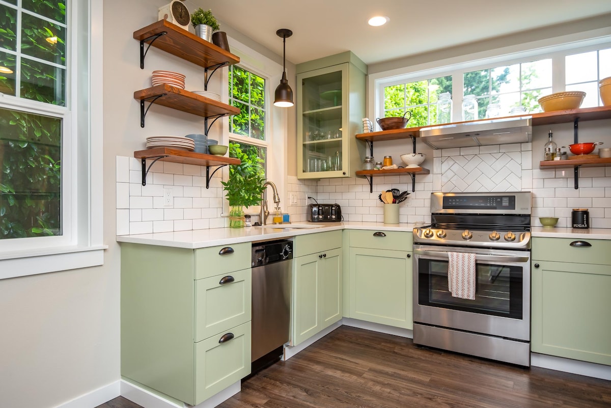 A modern kitchen with light green cabinetry is featured, complemented by open wooden shelving. Stainless steel appliances, including a gas stove, are neatly arranged. Natural light filters through large windows, illuminating the clean white backsplash and countertop.