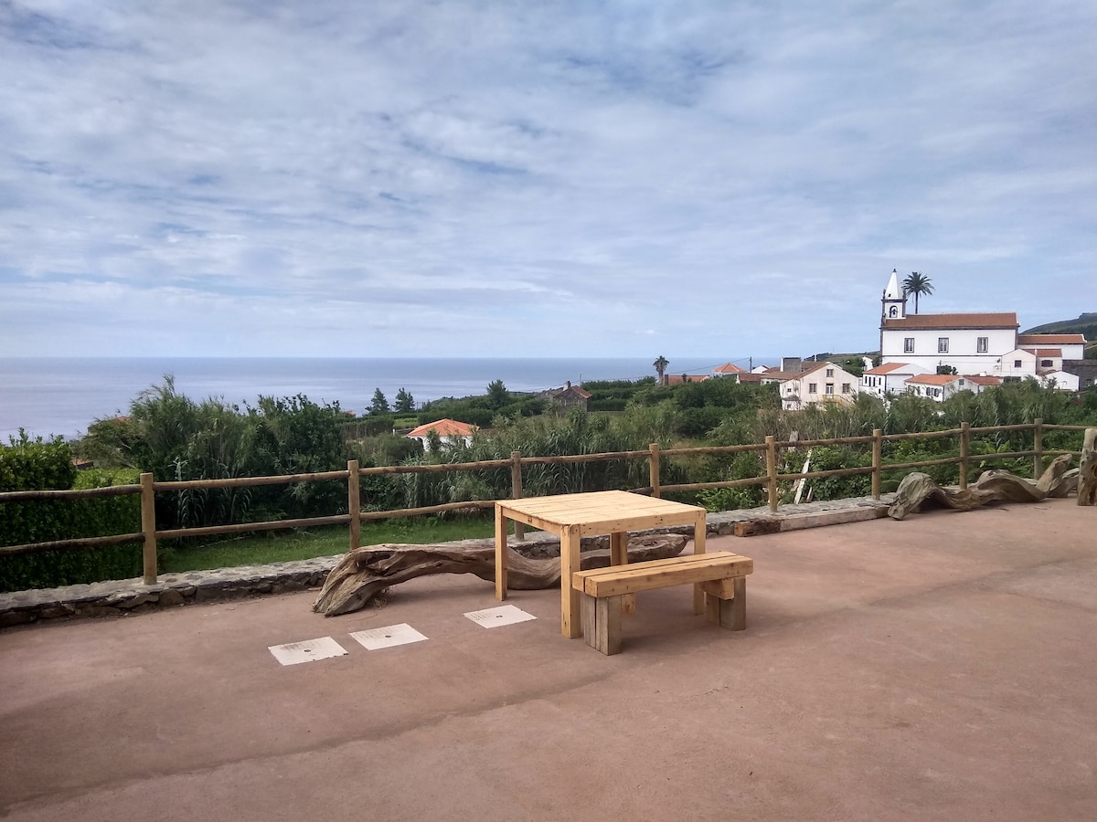 A wooden table and benches are positioned on the terrace, offering expansive views of the surrounding greenery and the ocean. In the background, traditional buildings and a church steeple are visible against a partly cloudy sky.