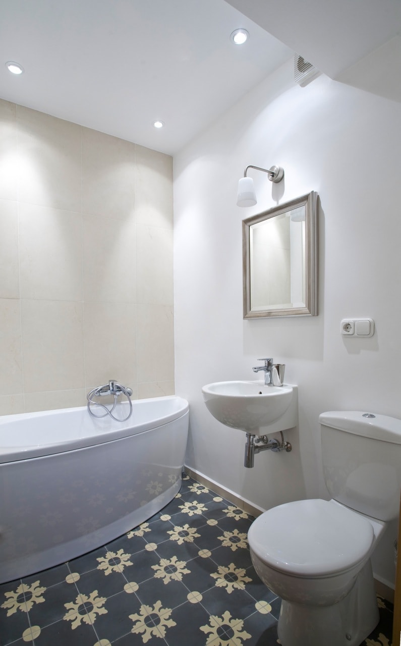 A compact bathroom is seen, featuring a curved bathtub alongside a sleek basin and wall-mounted faucet. A modern toilet is positioned nearby. Decorative tiles with a floral pattern cover the floor, while soft lighting is provided by recessed fixtures and a wall-mounted sconce.