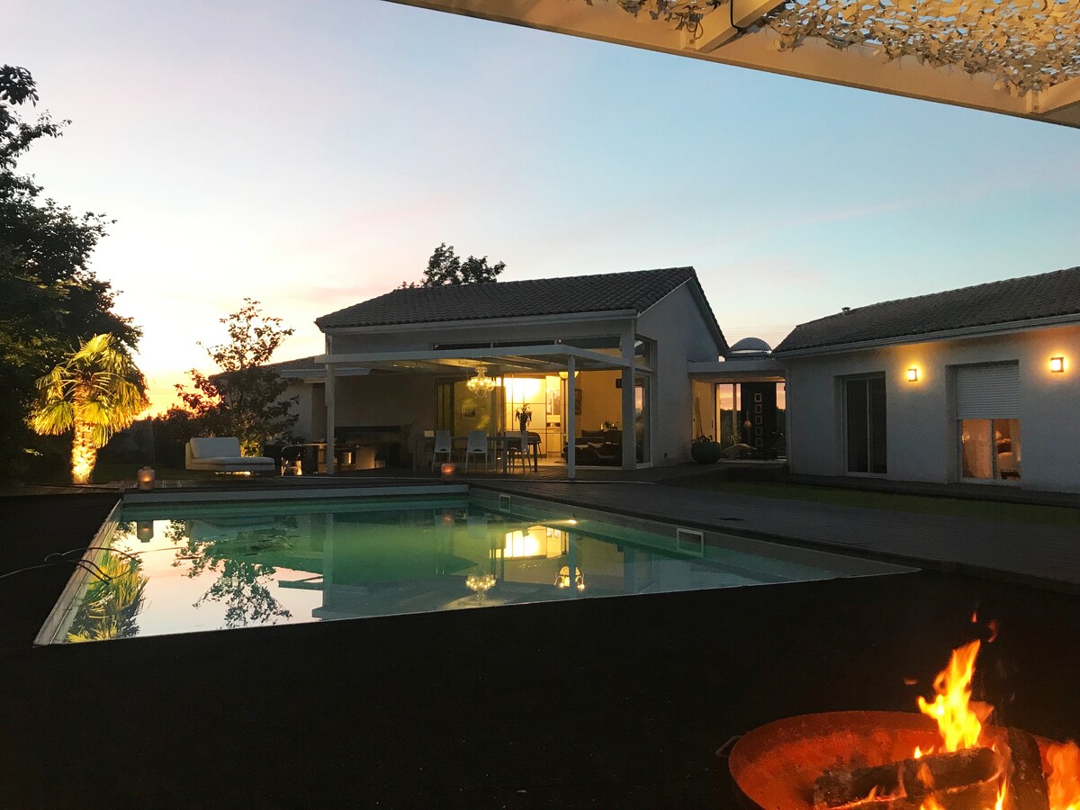 The image captures a modern villa at dusk, featuring a reflective swimming pool surrounded by landscaping and outdoor seating areas. A warm glow emanates from a fire pit in the foreground, complementing the soft colors of the sunset sky. Large glass doors open to living spaces.