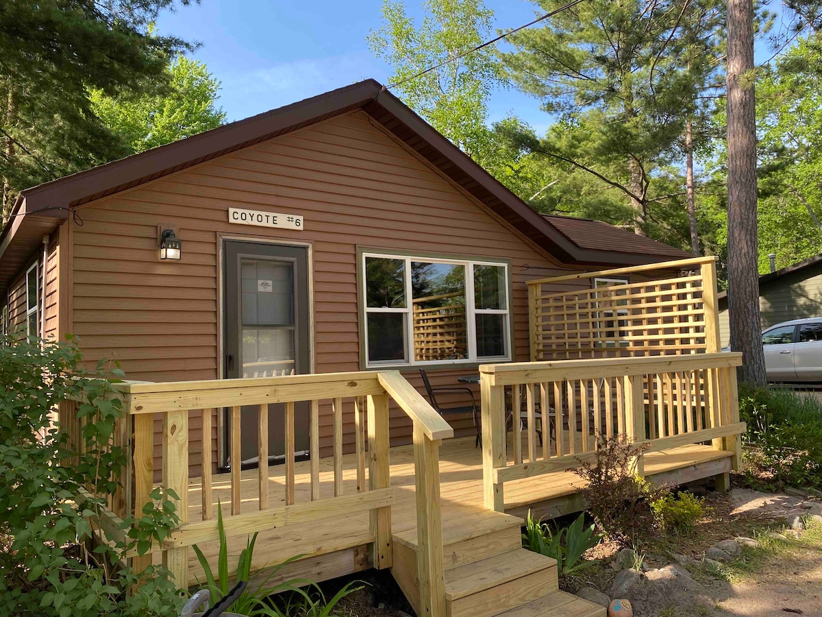 A charming cabin exterior presents a warm brown facade, framed by greenery. A welcoming front porch features a railing, with wooden steps leading up to the entrance. Large windows allow natural light to enter, providing views of the surrounding trees.