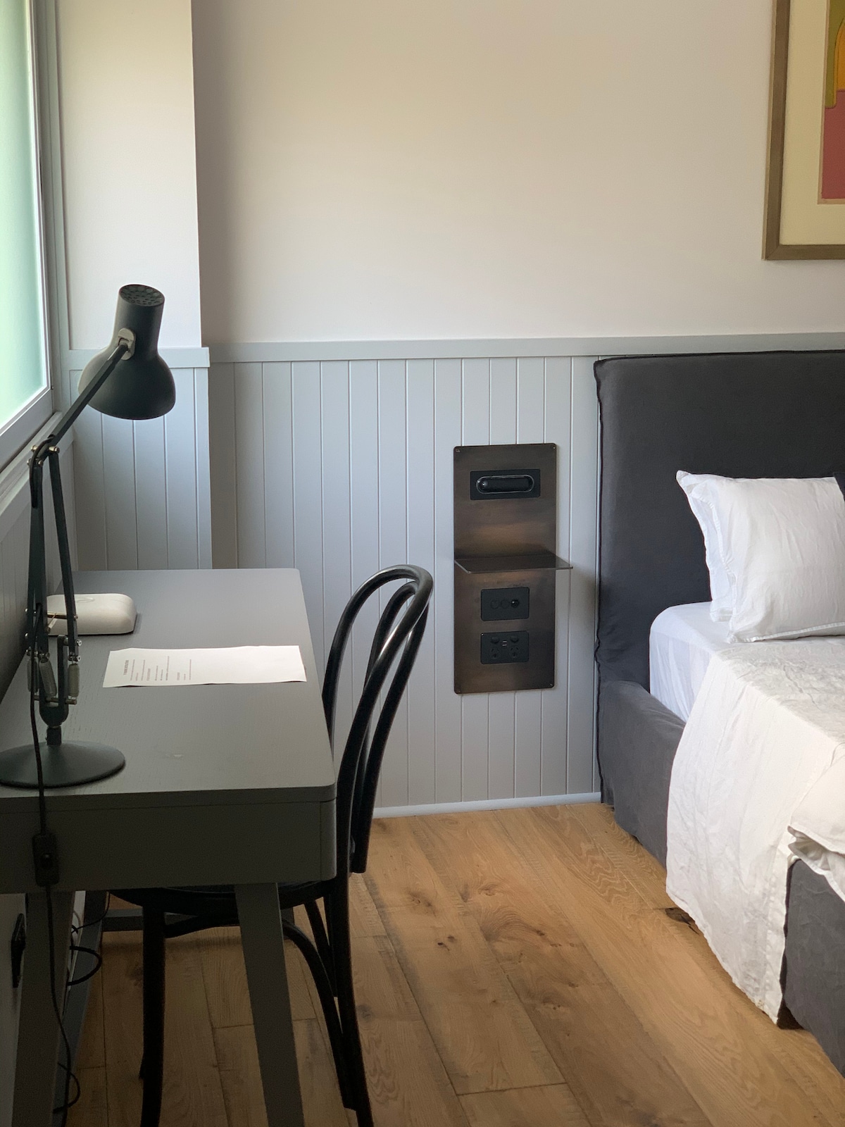 A well-lit corner of the studio features a simple writing desk with a lamp and a sheet of paper. Next to a spacious bed with crisp white bedding, a power outlet panel is mounted on the wall, providing easy access to charging points.