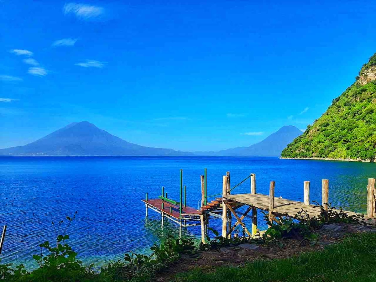 A private dock extends over clear blue waters, framed by mountains rising majestically in the distance. Lush greenery lines the shoreline, creating a serene landscape. The vibrant colors of the sky reflect on the lake's surface, enhancing the tranquil setting.