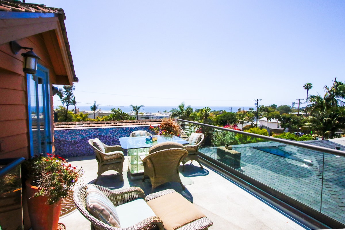 A rooftop terrace with a glass railing offers views of the ocean and surrounding landscape. Comfortable seating arrangements, including lounge chairs and a dining table, are arranged under a clear sky. Lush greenery and colorful flowers are visible in pots, enhancing the outdoor space.