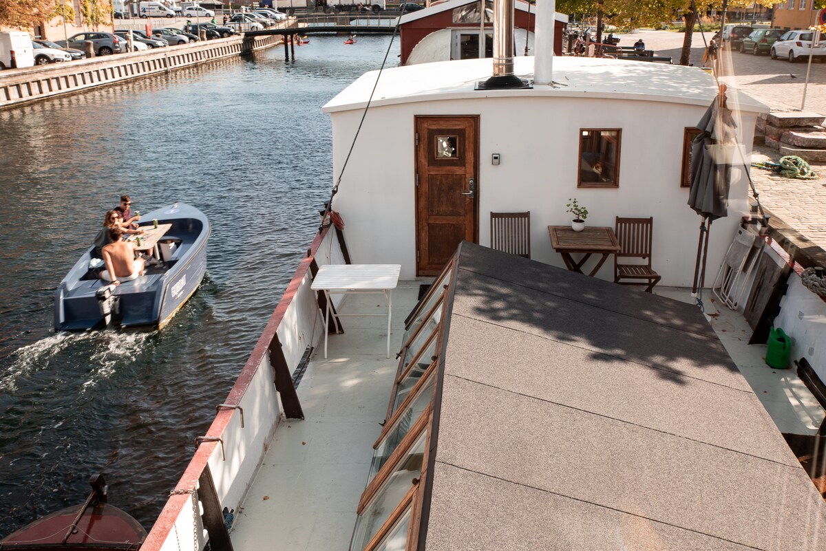 The image showcases the upper deck of the houseboat, featuring a sun deck with outdoor seating and a small table. A scenic waterway is visible nearby, with a boat passing by. The entrance door is prominently displayed, framed by the cozy exterior.
