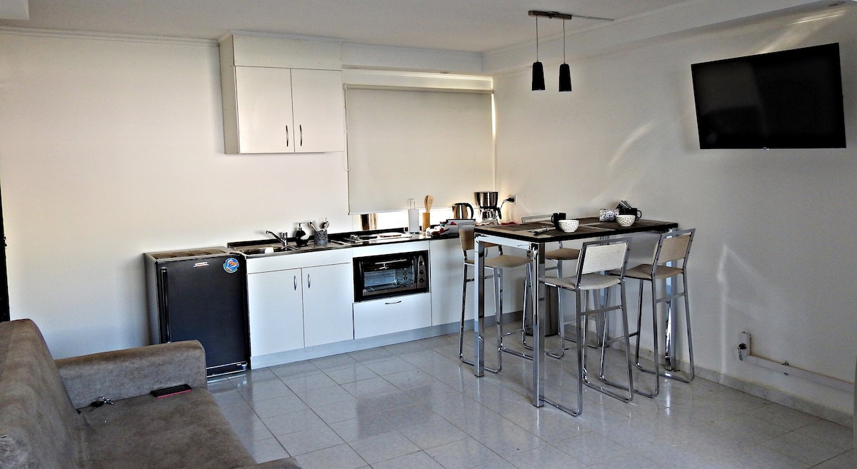 The image displays a modern kitchen area with sleek white cabinetry, equipped with essential appliances including a refrigerator and an oven. A dining table with four metal chairs is positioned nearby, and a wall-mounted television is visible, contributing to the open and functional layout.