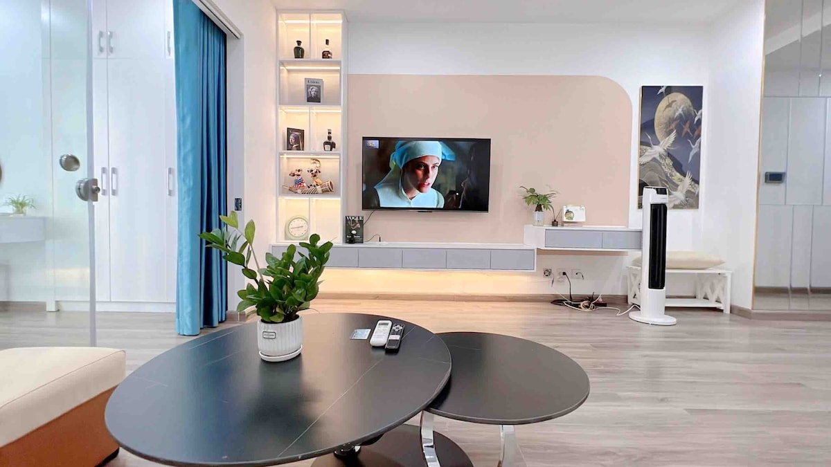 A modern living area features a sleek entertainment unit with a large TV mounted on the wall. Decorative shelves are illuminated, showcasing thoughtful decor. A green plant sits on a round table, contributing to the space's contemporary feel, while large windows allow natural light to fill the room.