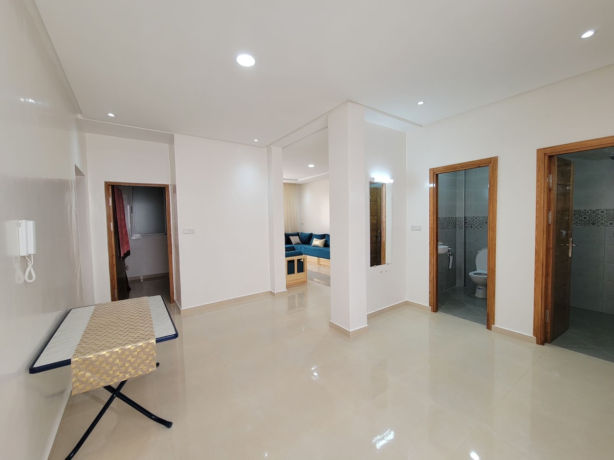 A spacious interior area features a glossy tiled floor and bright, neutral walls. Two doorways lead to bathrooms, while a large window allows natural light to fill the space. A folding ironing board stands against one wall, providing practical convenience.