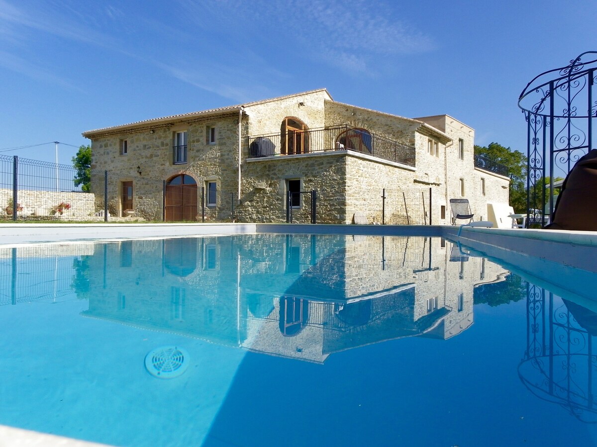 A stone building from the 17th century is reflected in a clear, tranquil swimming pool. The structure includes various balconies and large windows, showcasing the blend of historic charm and modern amenities. A clear blue sky can be seen above, providing a serene backdrop.
