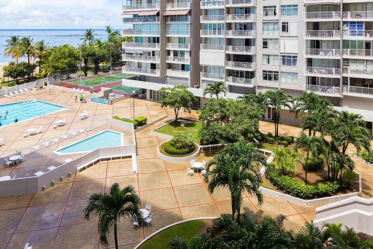 A spacious outdoor area is visible, featuring a large pool and landscaped gardens. Comfortable lounge chairs are placed around the pool, along with pathways bordered by palm trees and greenery. The ocean can be seen in the background, enhancing the tranquil atmosphere.