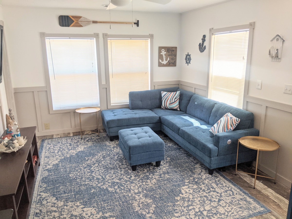 A spacious living room is furnished with a blue sectional couch and a matching ottoman. Two large windows provide natural light, adorned with blinds for privacy. A patterned area rug covers the floor, complementing a light wooden TV stand and coastal-themed decor on the walls.