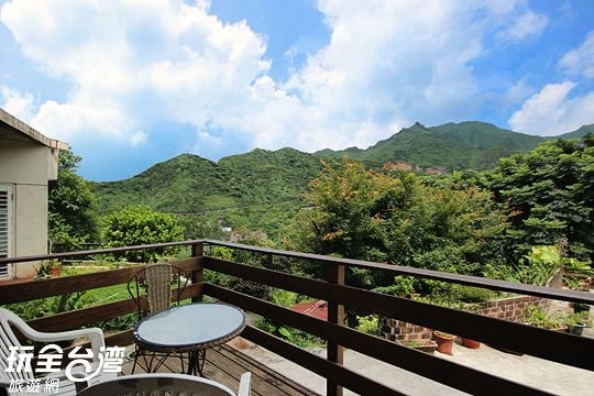 A wooden balcony offers a view of lush green mountains under a partly cloudy sky. A small round table is accompanied by a single chair, allowing for a peaceful outdoor seating area. Thickets of trees provide a natural border, enhancing the serene setting.