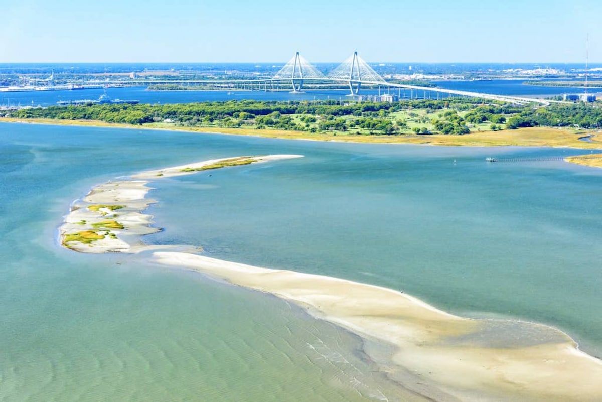 An aerial view captures the serene landscape of a coastal area, featuring a sandy shore and gentle waves. The iconic bridge spans across the water, connecting the landscape to the vibrant city beyond. Green landscapes and various waterways create a tranquil atmosphere.