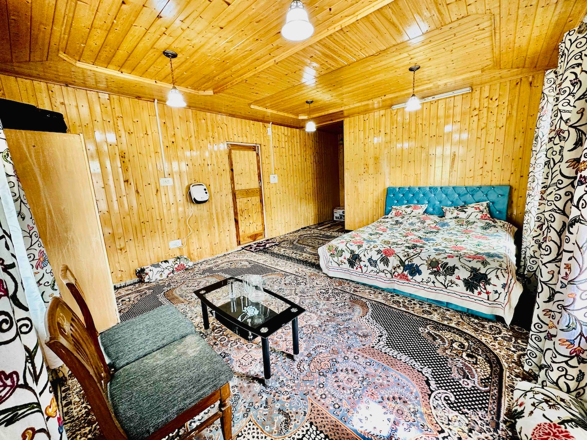 A spacious room features wooden paneling and a decorative carpet. A large bed is adorned with a colorful quilt. A small table is set in front of two chairs. Soft lighting is provided by ceiling fixtures, creating an inviting ambiance.