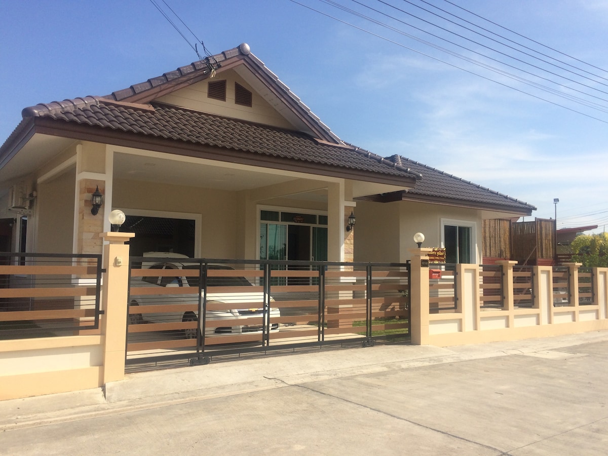 A single-story villa is featured, characterized by a tiled roof and light-colored exterior walls. A gated entrance provides access to a driveway, with a vehicle visible in front. Large windows enhance the facade, allowing natural light to illuminate the interior.