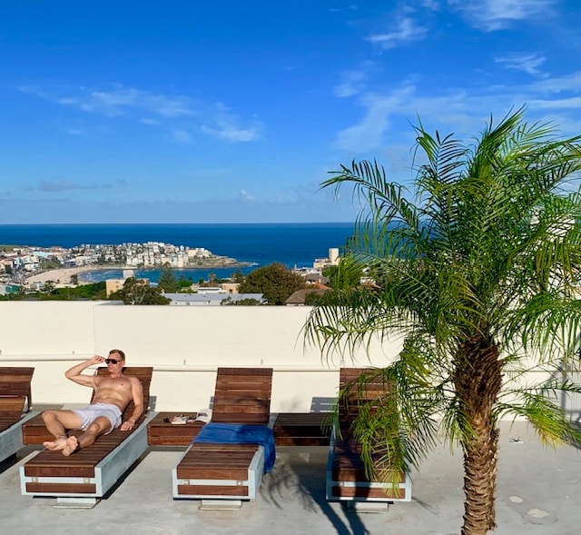 A rooftop area features several lounge chairs arranged under a clear blue sky. A palm tree provides a touch of greenery, while expansive views of the ocean and coastline can be seen in the background. The space invites relaxation in a sun-drenched setting.