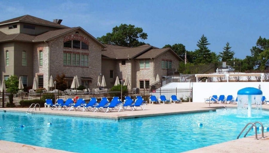 A large outdoor swimming pool is surrounded by tall, white fencing. Multiple lounge chairs in blue are arranged neatly around the pool area. A shaded area with umbrellas provides seating options, while a water feature adds a playful touch. A multi-story building is visible in the background.