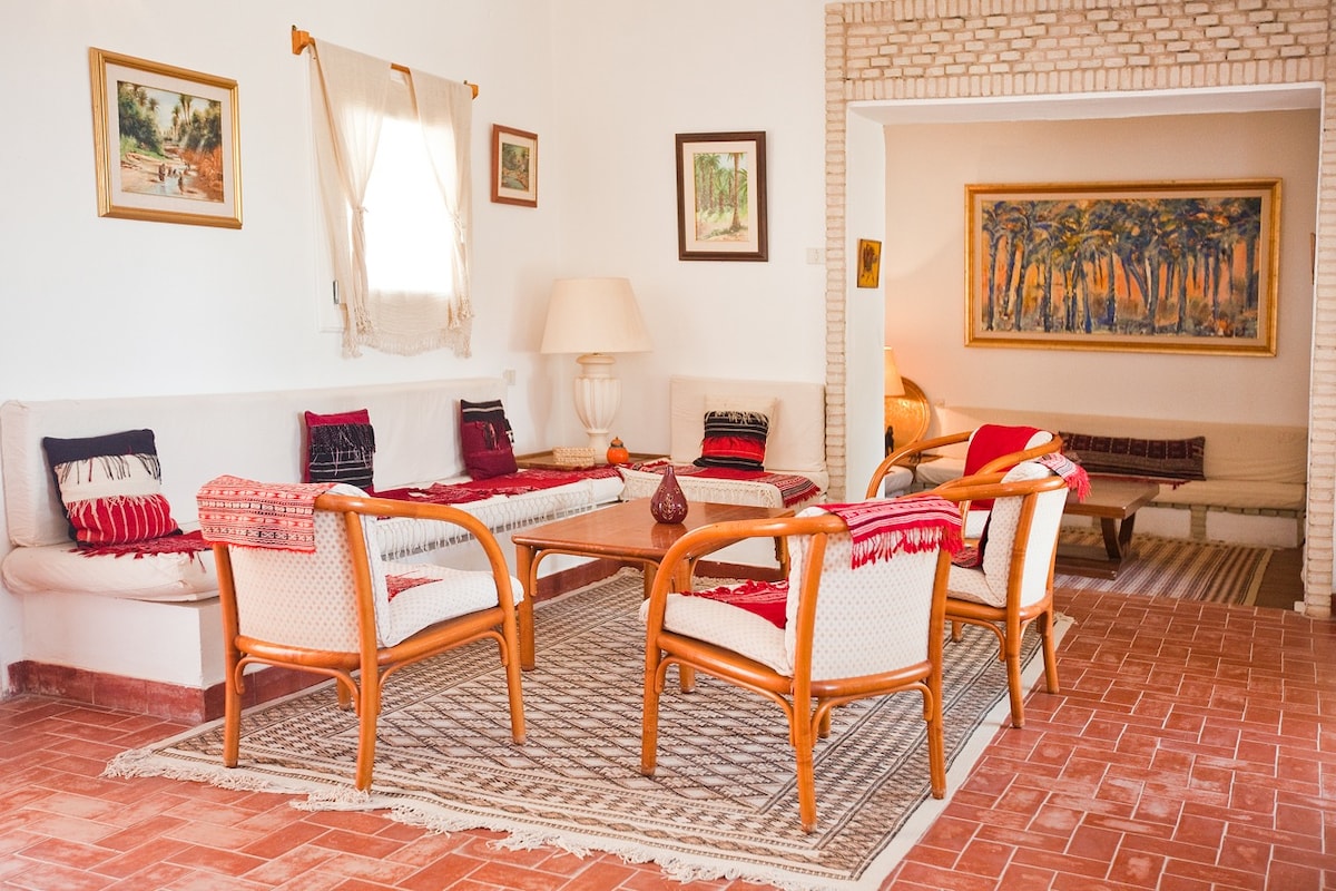 A spacious living area is arranged with comfortable seating, featuring woven chairs surrounded by decorative cushions. Warm light fills the room, accentuating the earthy tones of the walls and tiled flooring. Framed artworks adorn the walls, adding visual interest to the inviting space.