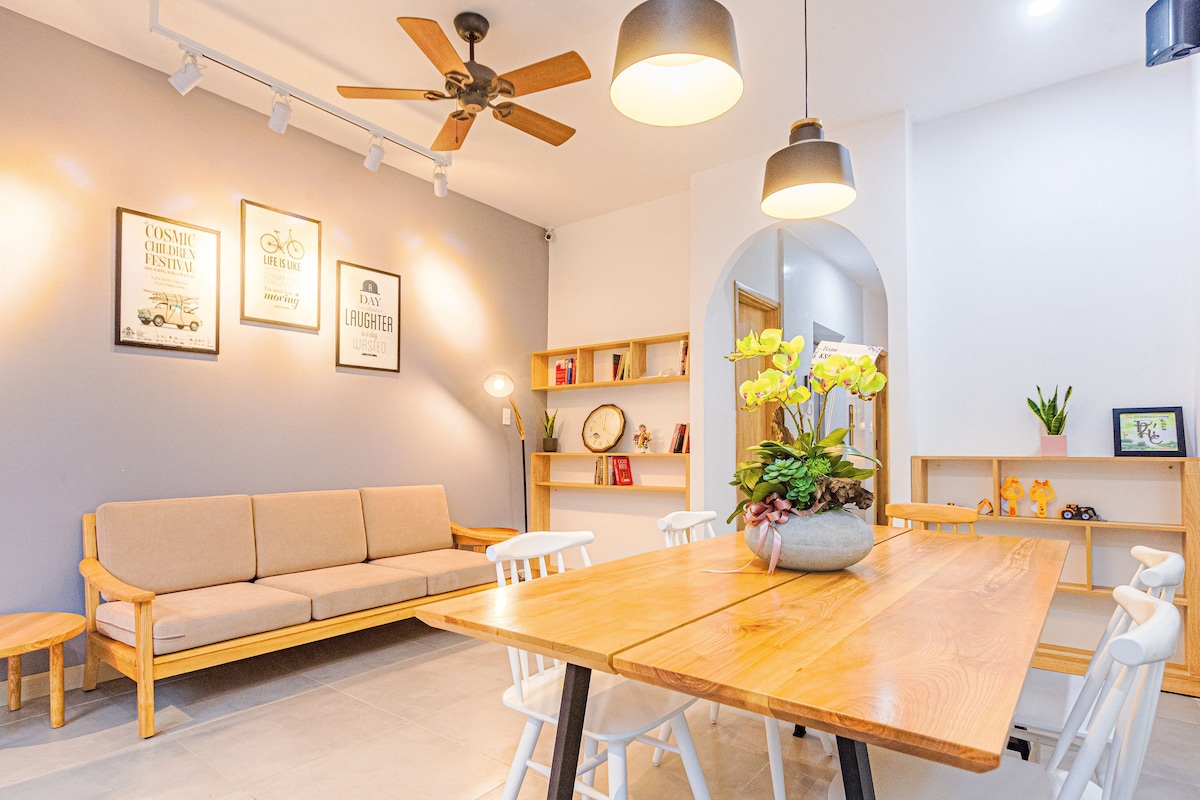 A spacious living area is presented with a wooden dining table and white chairs. A light-colored sofa is situated against a grey wall adorned with framed art. Natural light fills the space, highlighting a decorative plant at the center of the table.